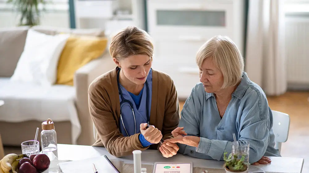 A healthcare professional in a brown cardigan and blue scrubs checks the hand of an older woman seated at a table in a bright, modern living room.