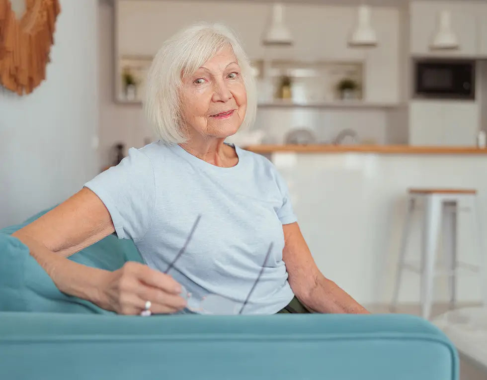 An older woman with short white hair sits on a teal sofa in a bright, modern living room, wearing a light blue t-shirt and holding a pair of eyeglasses.